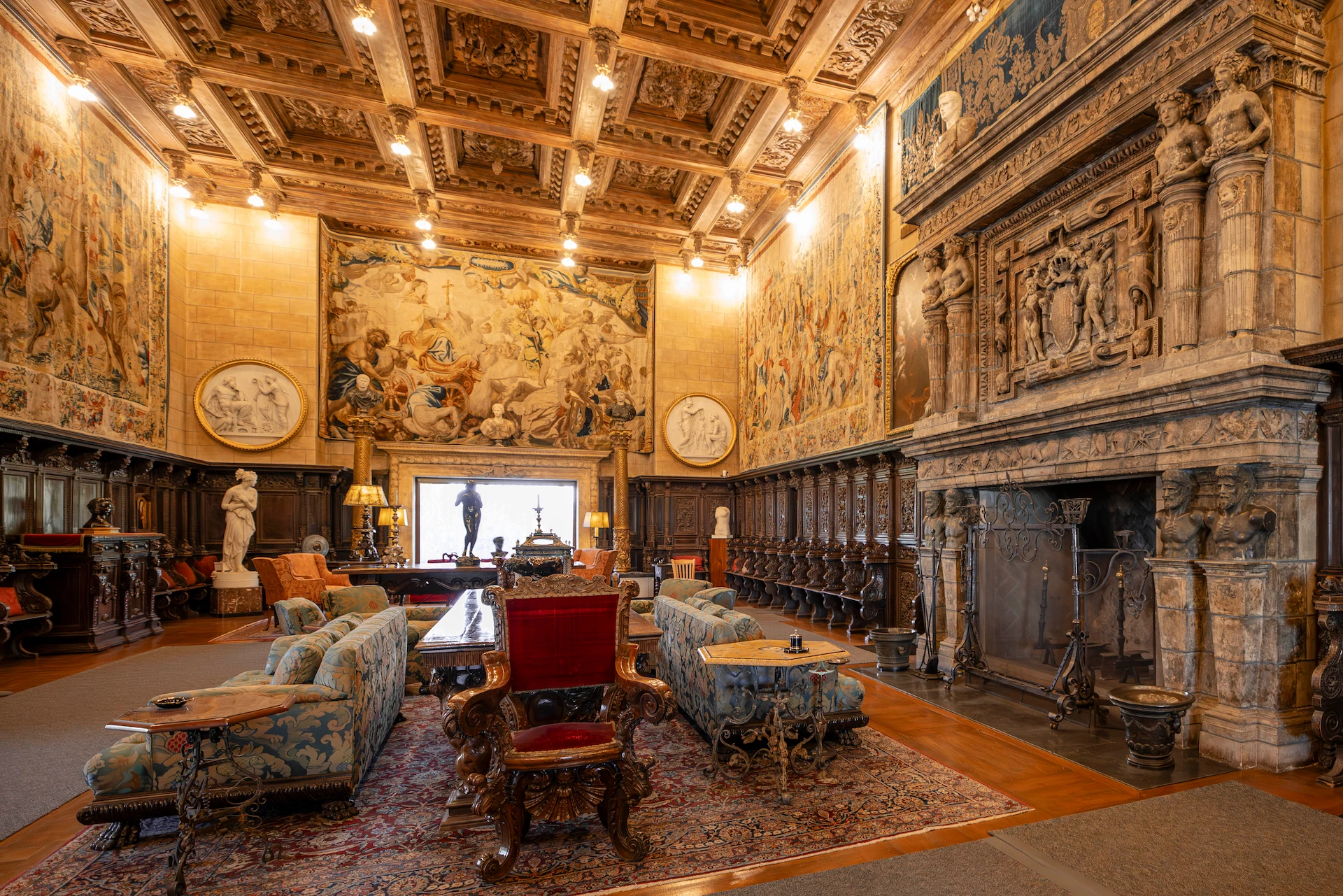 Luxurious historic lounge with carved wooden ceiling, tapestries, and ornate stone fireplace, filled with upholstered seating and tables.