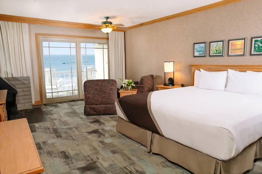 Hotel room with a king bed, nightstands, and a sliding glass door to a balcony overlooking the ocean.