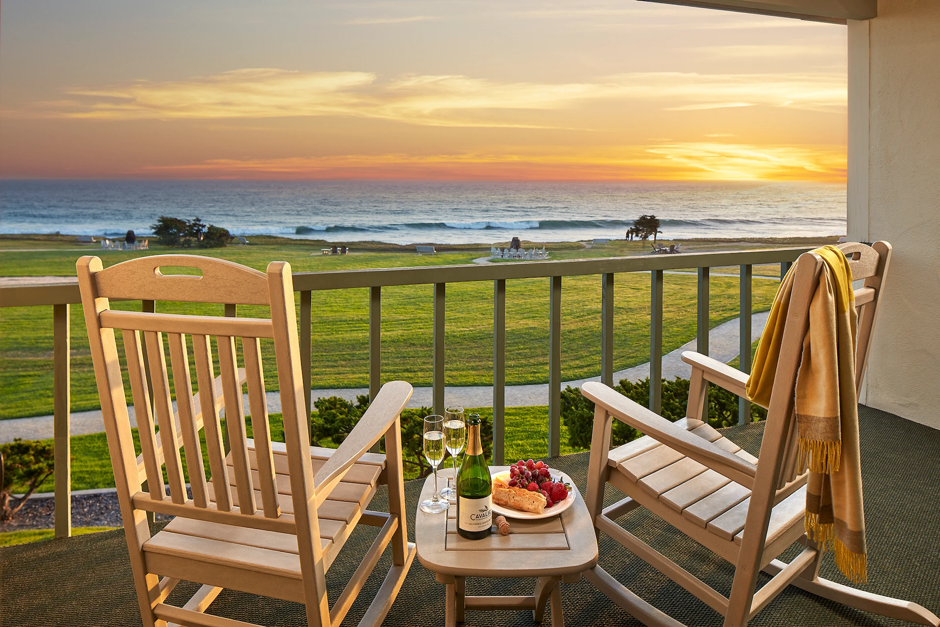 Balcony view at sunset with wooden chairs and a small table set with wine, glasses, grapes, and bread overlooking the ocean.