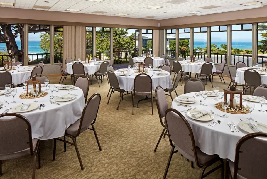 Banquet hall with round tables set for a formal dinner, white tablecloths, glassware, and an ocean view through large windows.