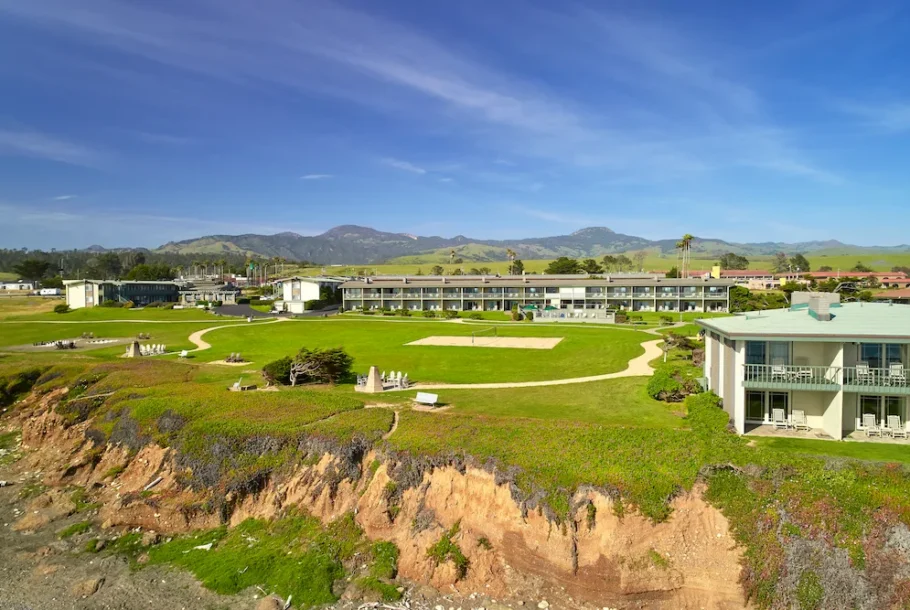 Coastal resort with green lawn, winding paths, and row of buildings against a backdrop of hills and blue sky.