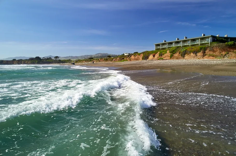 Sunny beach with turquoise water and white foam washing onto a sandy shore, a red cliff, and a long building atop the cliff in the background.