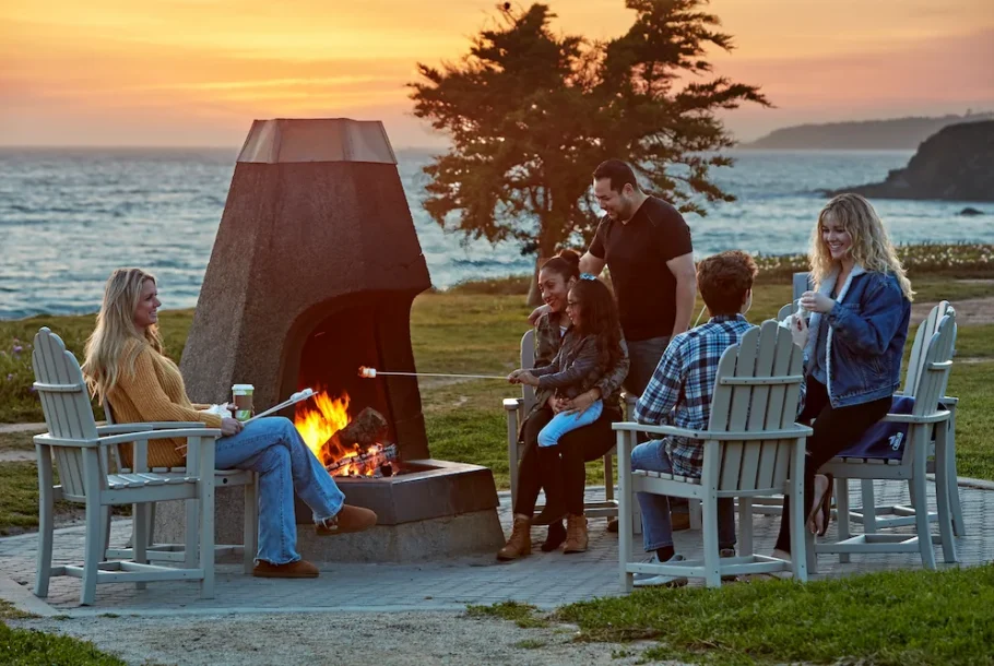Six friends seated in Adirondack chairs around a lit outdoor fire pit by the ocean at sunset, roasting marshmallows and chatting.