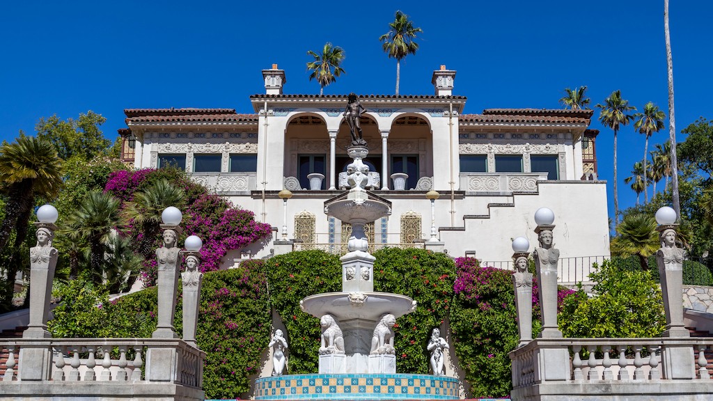Elegant white Mediterranean villa with an arched balcony, fountain centerpiece, and lush greenery.