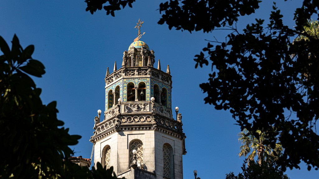 Ornate stone bell tower with arches and a colored dome, set against a clear blue sky and framed by tree branches.