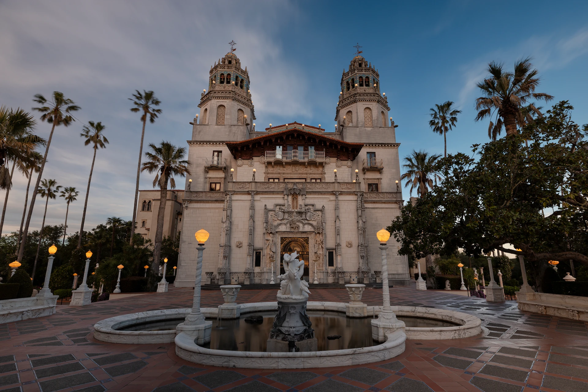 Front view of a grand historic building with two ornate towers, intricate stonework, and a circular fountain at the entrance, palm trees framing the scene at dusk.
