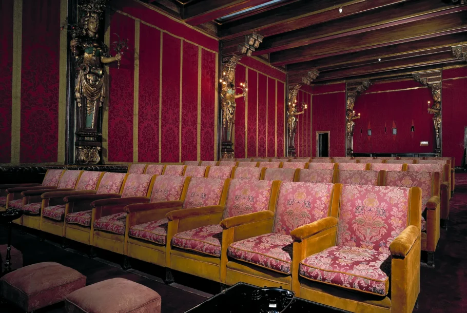 Interior of a vintage theater with red damask walls, gold trim, and pink upholstered seats awaiting audience.