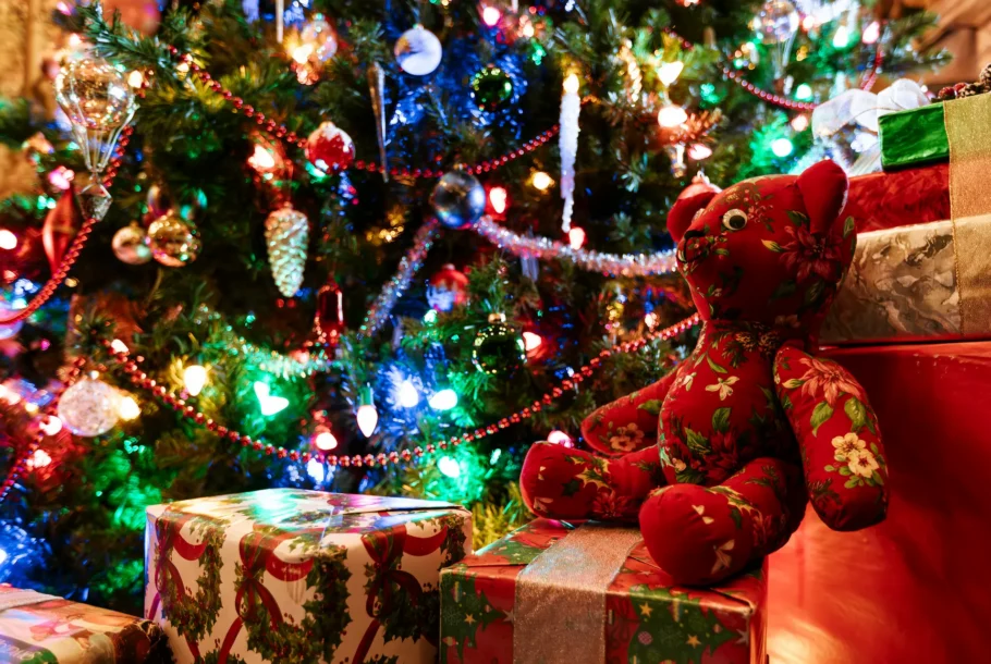 Colorful Christmas tree with baubles, lights, tinsel, and a red floral stuffed bear beside wrapped gifts.