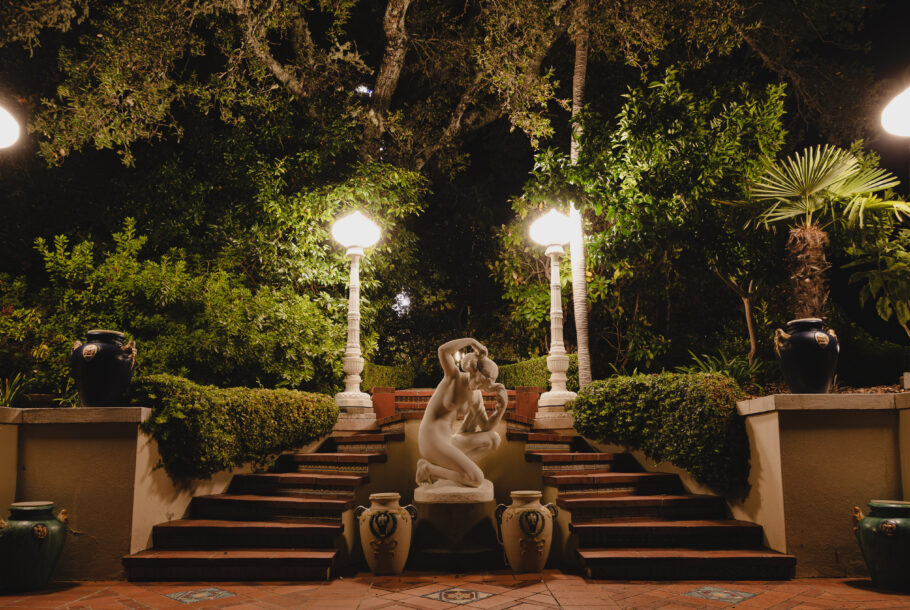 Nighttime garden courtyard with four glowing lamp posts, a classical statue group on steps, and lush green trees in the background.