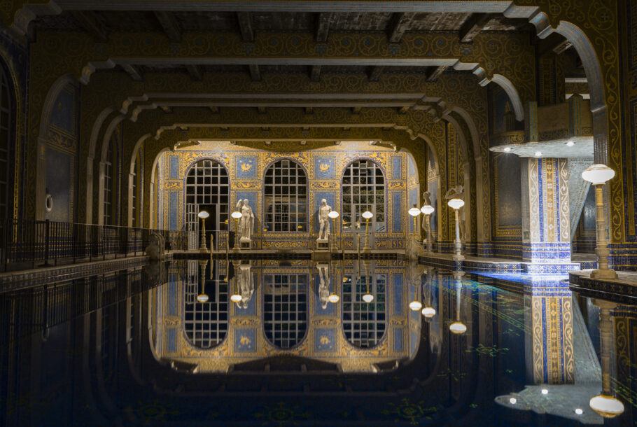 Grand, ornate indoor pool hall with blue-gold mosaics, arches, and statues reflected in still water at night, lanterns glowing.