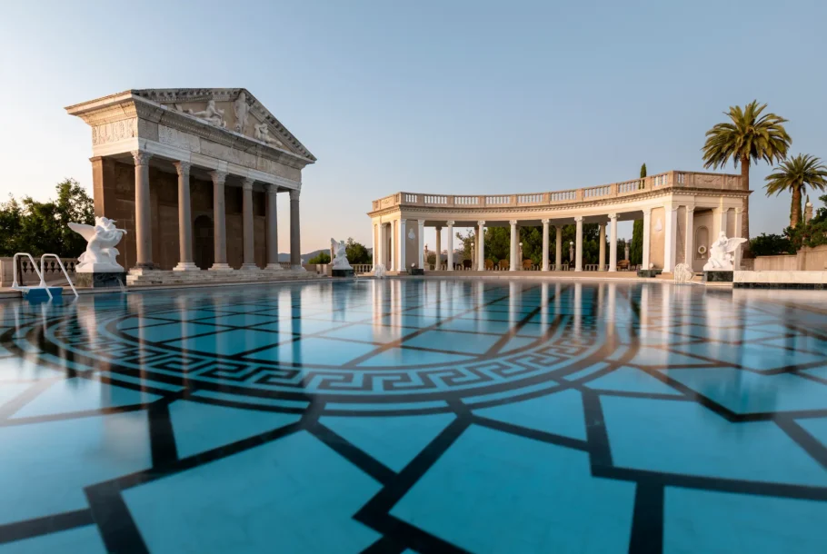 Classical temple and colonnade by a turquoise tiled pool with statues and palm trees in a sunny outdoor setting