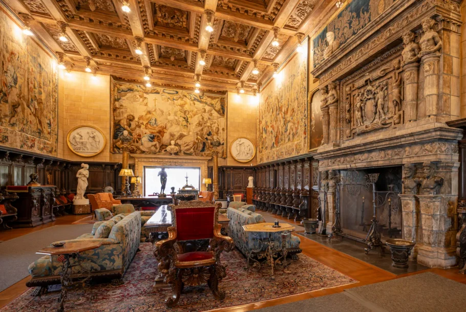 Luxurious historic living room with carved wooden ceiling, tapestries, and stone fireplace with statues on the right wall.